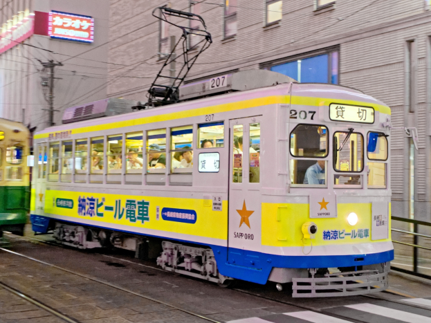 Eine gelbe und blaue Straßenbahn auf einer Stadtstraße bei Nacht mit Menschen darin, Gebäuden mit Fenstern, Strommasten mit Drähten und Schildern im Hintergrund.