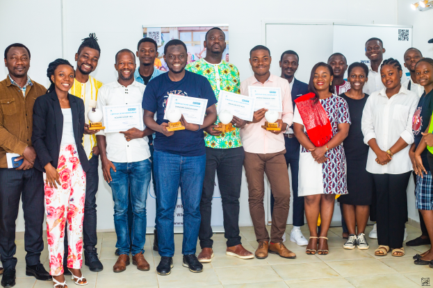 Eine Gruppe strahlender Menschen hält Zertifikate in einem Raum, mit einem Tisch voller Gegenstände rechts und Tafeln und einem Plakat im Hintergrund, feiert die WIK Awards 2019.