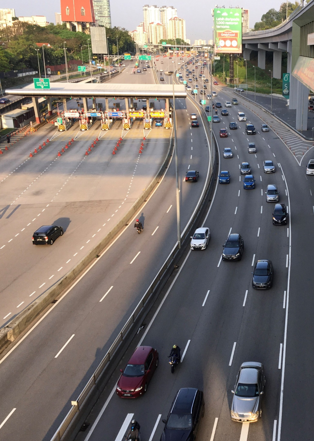 Eine befahrene Autobahn mit Autos, Motorrädern, Verkehrskegeln, Hinweisschildern, Laternen, einer Brücke, Bäumen, Gebäuden und einem klaren blauen Himmel.