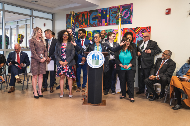 Eine Gruppe von Menschen, die in einem Raum stehen und sitzen, mit einem Rednerpult, einem Mikrofon und einer Tafel mit der Aufschrift "Sancho-Stadtrat tritt dem Stadtrat bei", mit Flaggen, einem Feuerlöscher und Deckenleuchten an der Rückwand.