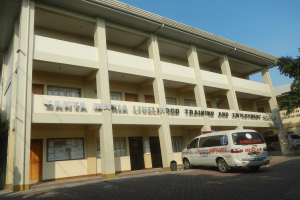 Außenansicht des Santa Maria Lebensmittel-Trainings- und Beschäftigungszentrums mit Fenstern, Türen und Beschilderung, umgeben von Bäumen und Fahrzeugen auf einer Straße unter einem klaren blauen Himmel.