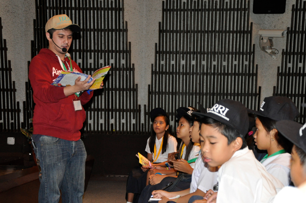 Ein Mann in einer rotbraunen Kapuzenjacke und Jeans hält ein Buch und spricht in ein Mikrofon, mit ein paar Kindern in weißen T-Shirts und schwarzen Mützen auf der rechten Seite und einer Überwachungskamera an der Wand im Hintergrund.