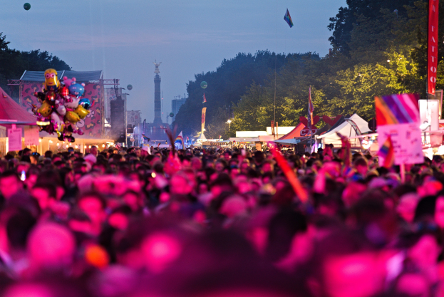 Eine gro├če Menge Menschen auf einem nächtlichen Musikfestival, beleuchtet von bunten Lichtern, umgeben von Zelten, Fahnen, Masten und anderen Gegenständen, mit einem Turm, Bäumen und einem klaren Nachthimmel im Hintergrund.