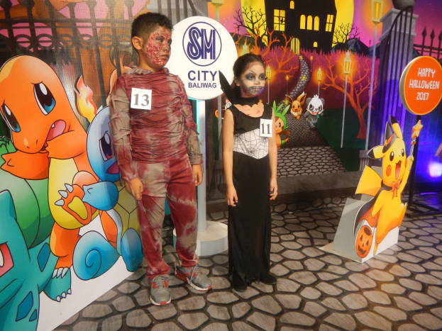 Zwei Kinder in Halloween-Kostümen stehen nebeneinander vor einer Wand mit einer Gemälde von einem Gebäude, Bäumen, Laternenmasten und einem Zaun, mit Schildern mit Text und Tierbildern und Festbeleuchtung.