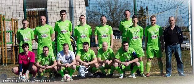 Eine Gruppe von Männern in grünen T-Shirts steht auf einem Fußballfeld, einige sind in der Hocke, mit einem Ball und einem Torpfosten im Blickfeld; der Hintergrund umfasst Bäume, Gebäude und Fahrzeuge.