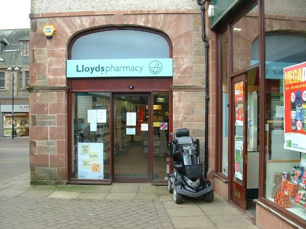 Eine Apotheken-Filiale mit einem vor ihr geparkten Fahrzeug und einem Gebäude in der linken Ecke.