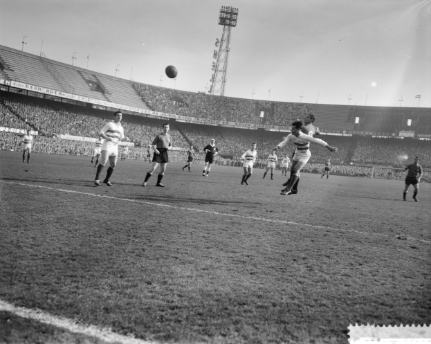 Eine Gruppe von Männern, die auf einem Feld Fütball spielen, mit einem Stadion, Flutlicht und einem klaren Himmel im Hintergrund.