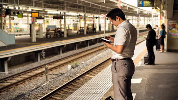 Ein Mann steht auf einem Bahnsteig und schaut auf sein Handy, umgeben von anderen Menschen, mit Bahnschienen und einer Tafel mit Text im Hintergrund.