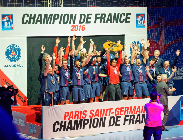 Eine Gruppe von Männern hält einen Pokal auf der Bühne, während eine Videokamera aufnimmt und ein "Champion de France 2016"-Plakat im Hintergrund zu sehen ist.