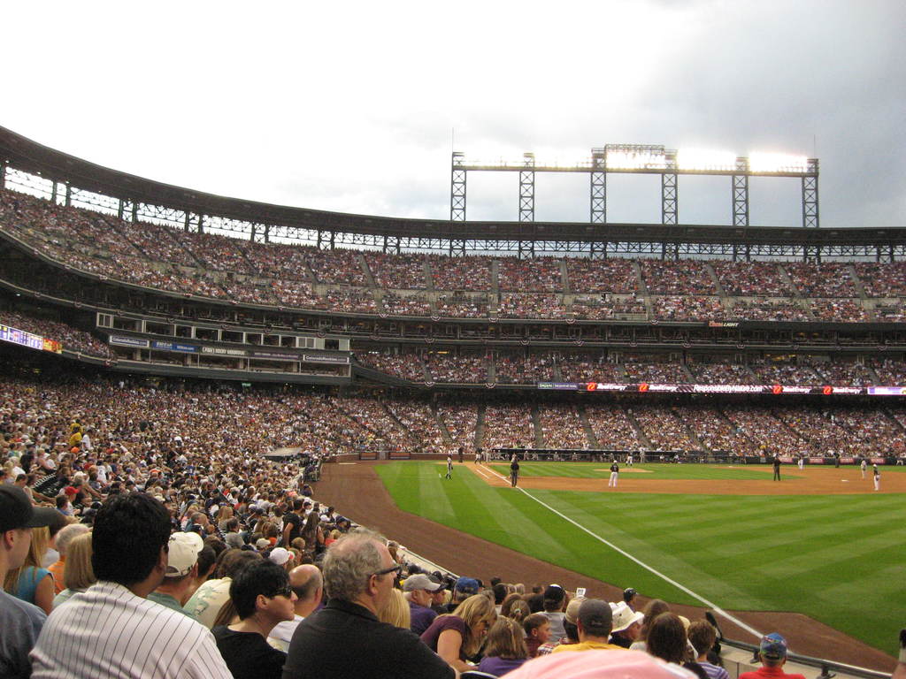 Ein Cricket-Stadion mit Spielern auf dem Feld und Zuschauern in den Rängen unter einem Metallrahmen mit Scheinwerfern.