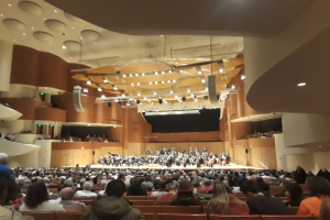 Eine große Halle voller Menschen, die ein Konzert genießen, mit Musikern auf der Bühne und anderen im Publikum, unter einer mit Lichtern geschmückten Decke.