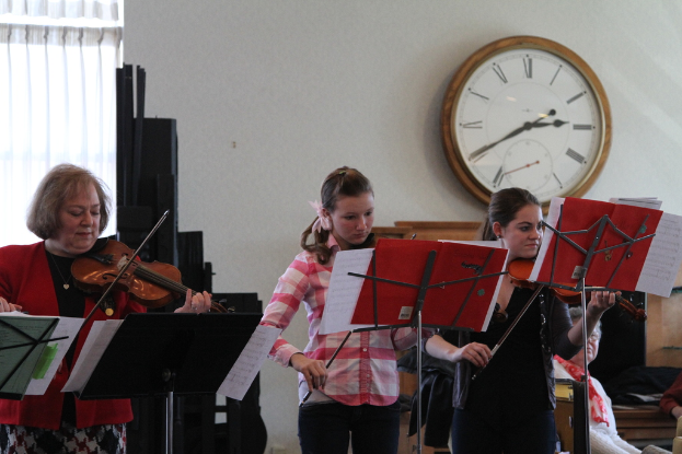 Drei Frauen auf der Bühne mit Geigen stehend und Notenpulte vor sich, während eine Gruppe sitzender Frauen im Vordergrund zuhört; eine Uhr hängt an der weißen Wand im Hintergrund, mit einem weißen Vorhang in der linken Ecke.