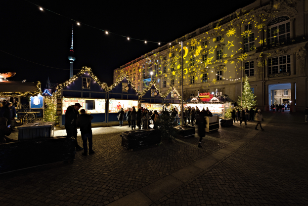 Ein lebendiger Weihnachtsmarkt in Berlin, Deutschland, mit Menschen um hell erleuchtete Stände, festliche Deko und Gebäude mit Fenstern im Hintergrund unter einem dunklen Himmel.