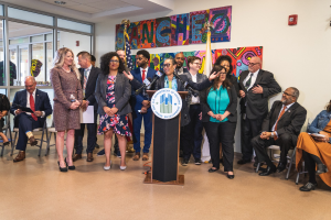Gruppe von Menschen in einem Raum mit einem Podium, einem Mikrofon und einer Tafel mit der Aufschrift "Sancho-Stadtrat tritt dem Stadtrat bei", umgeben von Flaggen, Sitzgelegenheiten und einem Feuerlöscher.