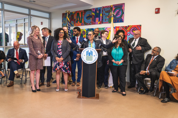 Gruppe von Menschen in einem Raum mit einem Podium, einem Mikrofon und einer Tafel mit der Aufschrift "Sancho-Stadtrat tritt dem Stadtrat bei", umgeben von Flaggen, Sitzgelegenheiten und einem Feuerlöscher.