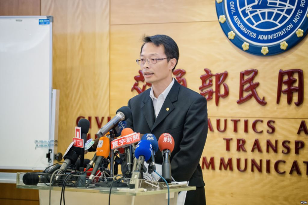 Ein Mann in einem schwarzen Blazer, weißem Hemd und schwarzen Hosen steht an einem Podium mit Mikrofonen, spricht auf einer Pressekonferenz mit einer hölzernen Wand mit Logo und Text dahinter und einem weißen Brett links von ihm.