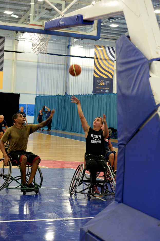 Eine Gruppe von Menschen in Rollstühlen spielt Basketball in einer Turnhalle mit einem Basketballkorb, Bannern mit Text und Deckenlampen.