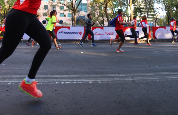 Eine Gruppe von Menschen, die bei einem Marathon auf einer Straße mit Bäumen, Gebäuden mit Fenstern, Bannern mit Text, Laternen und einem klaren blauen Himmel laufen, wobei einige Läufer Rucksäcke und Turnschuhe tragen.