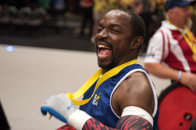 Ein Mann im Rollstuhl mit ausgestreckten Armen und einer Medaille um den Hals lächelt im Vordergrund, mit Menschen, Stühlen und anderen Gegenständen im Hintergrund.