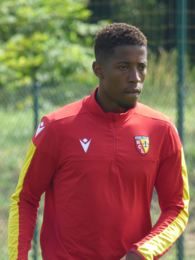 Ein junger Mann in einem roten und gelben Trikot steht auf einem Fußballfeld, das von einem Zaun und Bäumen umgeben ist und schaut entschlossen.