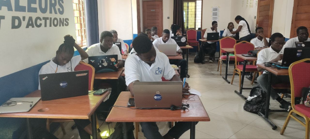 Eine Gruppe von Schülern in einem Klassenzimmer nutzt Laptops an Tischen, mit Taschen auf dem Boden und einer wandmontierten Textanzeige im Hintergrund, die auf einen Online-Kurs in Ghana hinweist.