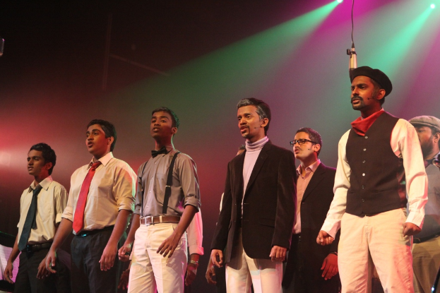 Eine Gruppe von Männern führt ein Drama auf der Bühne auf, im Hintergrund sind grüne Spotlights zu sehen.