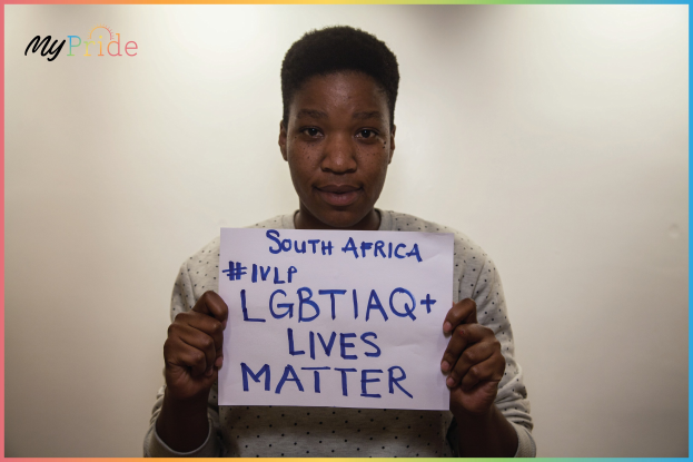 Eine Frau in einem weißen Hemd hält ein Schild hoch, auf dem in fetten schwarzen Buchstaben "LGBTQ+ Lives Matter" steht und steht entschlossen vor einer Wand.