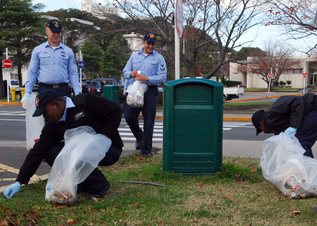 Drei Polizisten in Uniform sammeln M√∞ll an einer Stra√enseite, mit Bäumen, Gebäuden, Fahrzeugen und einem Mülleimer im Hintergrund.