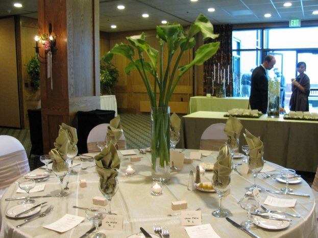 Ein Hotelrestaurant mit einer weiß gedeckten Tafel, die mit Tellern, Besteck und einem Glas gedeckt ist, umgeben von Stühlen, einem grünen Tisch und einem Mann in einem schwarzen Blazer und einer stehenden Frau dahinter, in der Nähe einer Tür, an der "Ausgang" steht und hinter der zwei Männer stehen.