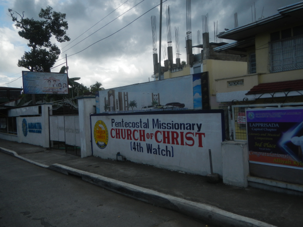 Die Pfingstmissionarische Kirche Christi 14. Wache steht mit einer Straße im Vordergrund, einer Wand mit Bannern und Text, Gebäuden, Bäumen, Pfählen, Drähten und einem bewölkten Himmel im Hintergrund.