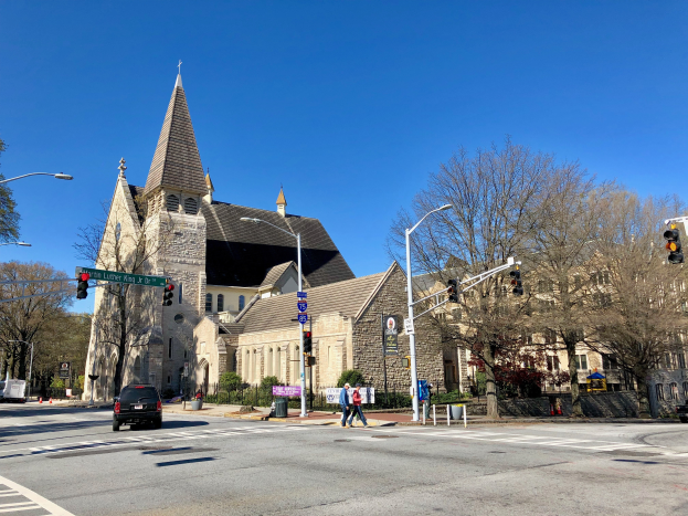 St. Lukes Episkopalkirche, eine große Kirche mit einem Turm, steht an einer Straßenecke umgeben von Gebäuden, Fahrzeugen, Fußgängern und Bäumen unter einem klaren blauen Himmel.