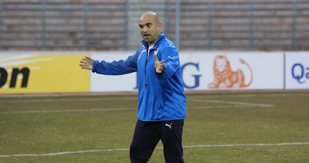 Ein Mann in einer blauen Jacke steht auf einem Fußballfeld, umgeben von Tafeln mit Text und Logos, entschlossen während eines Trainings.