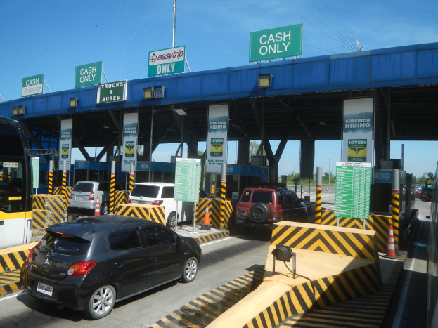 Eine Mautstation mit parkenden Autos davor, Fahrzeugen auf der Straße, Verkehrskegeln, Schildern mit Text, einer Brücke, Polen, Drähten, Bäumen und einem klaren blauen Himmel im Hintergrund.