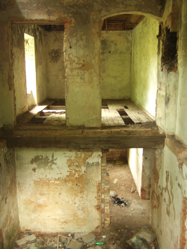 Ein alter Raum mit abblätiger Farbe, einem zentralen Fenster und verschiedenen Gegenständen auf dem Boden, die an einen Bombenschutzraum erinnern.