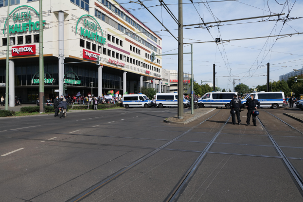 Eine Gruppe von Polizisten steht an der Straße neben einer Tram, mit Fahrzeugen, Strommasten, Gebäuden mit Namensschildern, Bäumen und einem bewölktem Himmel im Hintergrund.