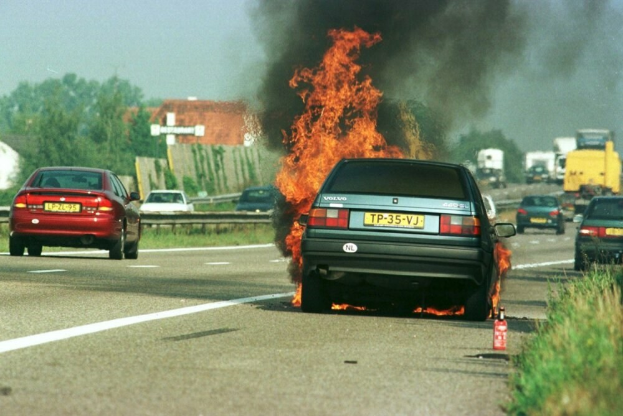 Ein Auto, das von Flammen umgeben ist, auf der Seite der Straße, umgeben von anderen Fahrzeugen, mit Bäumen, Gebäuden und einem klaren blauen Himmel im Hintergrund und einem Feuerlöscher auf der rechten Seite des Bildes.