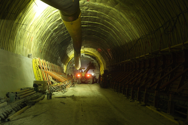 Eine Baustelle mit einem großen Tunnel in der Mitte, Fahrzeuge auf einer Straße, verstreute hölzerne Gegenstände, Rohre und eine Wand auf der linken Seite, beleuchtet von Deckenlampen.