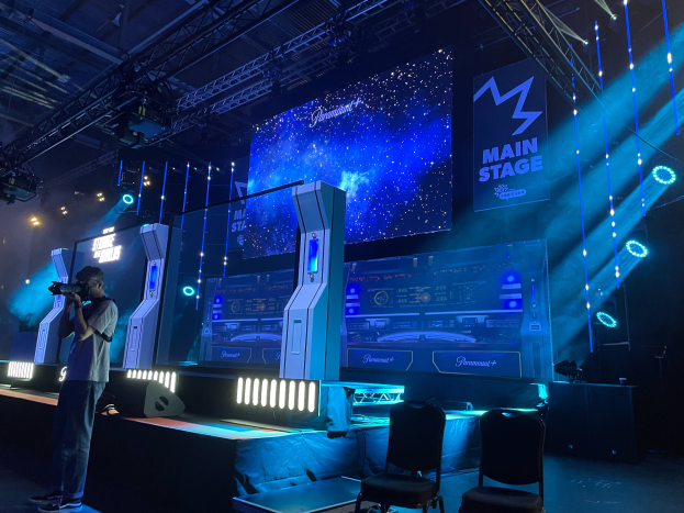 Ein Mann steht auf einer Bühne und hält eine Kamera, umgeben von Stühlen und anderen Gegenständen, sowie Bildschirmen und Lichtern im Hintergrund, was auf ein Gaming-Event hinweist.