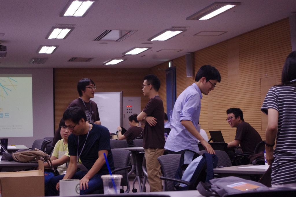 Ein Klassenzimmer mit Tischen, Stühlen, Laptops, Taschen, Menschen, die stehen und sprechen, einem Projektionsschirm, einer Holzwand und einer Tür im Hintergrund.