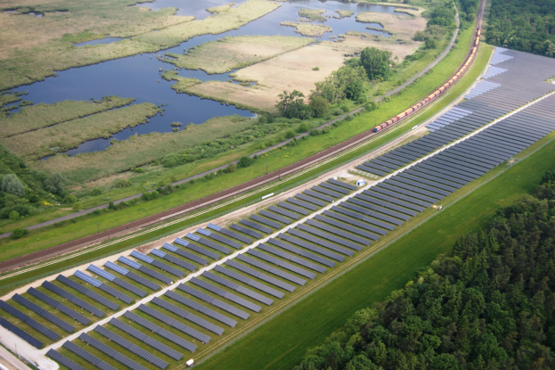 Luftaufnahme einer Solaranlage mit Solarpanelen auf einem Feld, umgeben von Bäumen, Gras, Pflanzen und Wasser, mit einem Zug auf einer nahen Bahnschiene.