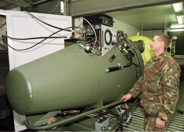 Ein Mann in militärischer Uniform steht neben einer großen grünen Bombe in einem unordentlichen industriellen Setting mit Maschinen, Drähten und einer beleuchteten Wand im Hintergrund, mit Text unten.