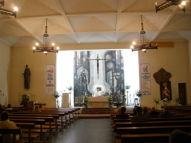 Eine Kircheninnenansicht mit Menschen auf Kirchenbänken, einer Person an einem Rednerpult mit Mikrofon, Kronleuchtern, Blumenbouquets, Kerzen und Plakaten an den Wänden.