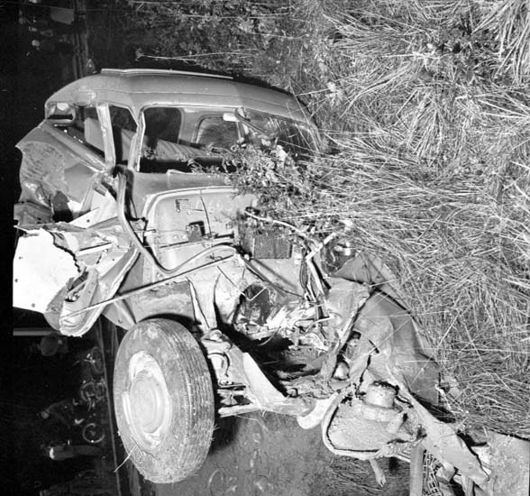 Schwarz-weiß-Foto eines schwer beschädigten Autos mit eingedrückter Front und zersplitterten Fenstern am Straßenrand, mit einem Baum und verstreuten Gegenständen im Hintergrund.