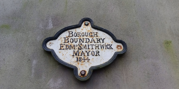 Schwarze Metalltafel mit weißer fettgedruckter Schrift, die "Gemarkungsgrenze Edm Smithwick Bürgermeister" lautet, an einem Gebäude angebracht.