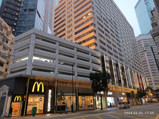 McDonald's-Restaurant auf einer belebten Stadtstraße mit hohen Gebäuden, Fahrzeugen, Passanten, Straßenmöblierung, Bäumen und einem klaren Himmel.