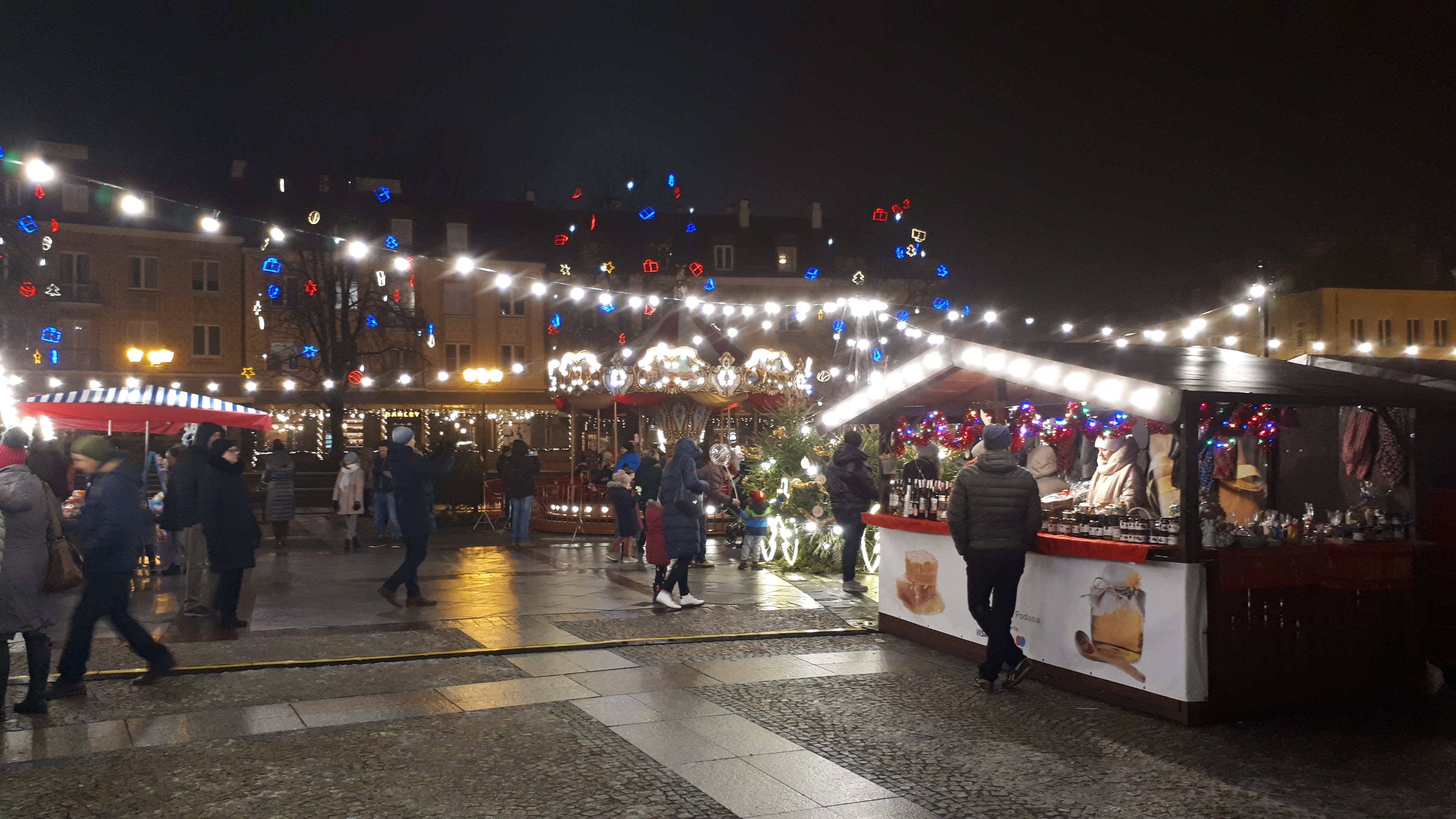 Ein belebter Weihnachtsmarkt in einer nächtlichen Stadt mit Menschen, Ständen mit Lichtern und Dekorationen, Gebäuden, Bäumen und festlicher Beleuchtung am Himmel.