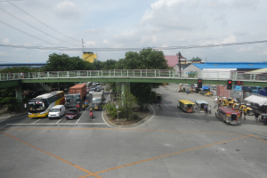 Eine belebte Straße mit Autos, Bussen und Fußgängern, eine Brücke mit Geländer, Ampeln, Gebäude, Bäume, Strommasten mit Kabeln und ein bewölkter Himmel.