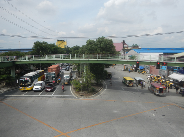 Eine belebte Straße mit Autos, Bussen und Fußgängern, eine Brücke mit Geländer, Ampeln, Gebäude, Bäume, Strommasten mit Kabeln und ein bewölkter Himmel.