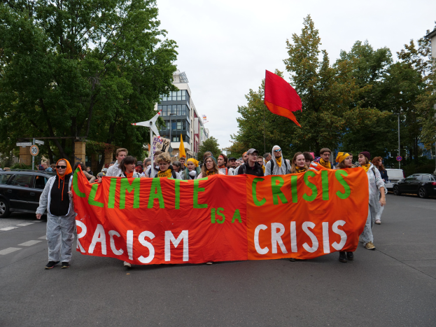 Eine Gruppe von Menschen marschiert eine baumbestandene Straße entlang und hält ein Transparent mit der Aufschrift "Klima-Krise ist eine Krise" in den Händen, mit Gebäuden und Fahrzeugen im Hintergrund.
