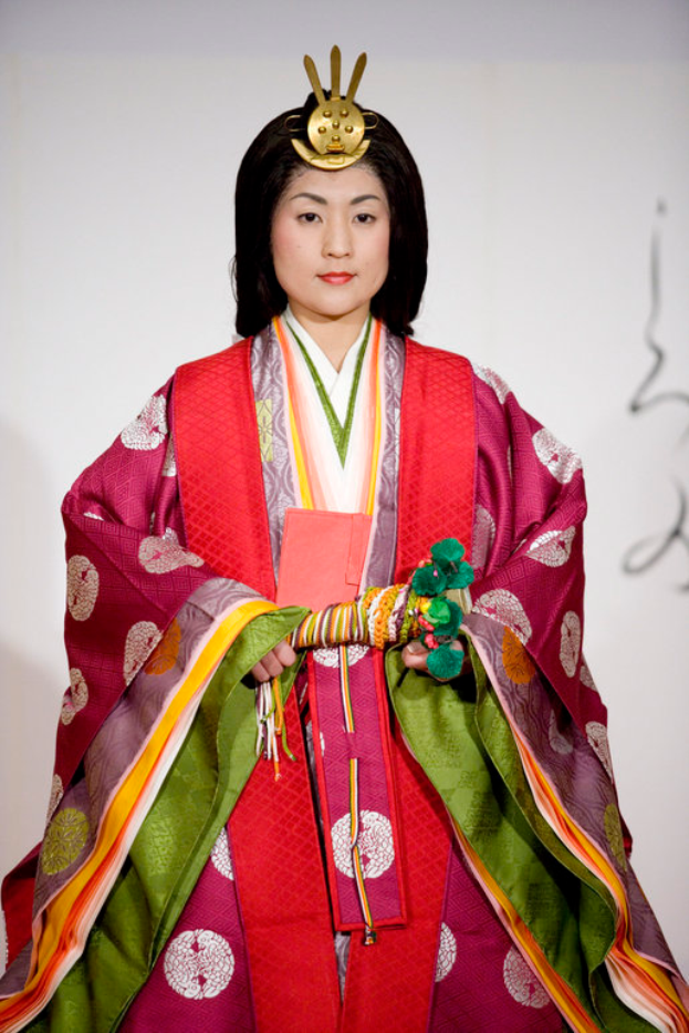 Eine Frau in einem traditionellen japanischen Kimono, das ein leuchtend rotes und grünes Kleid mit kunstvollen Mustern trägt, steht selbstbewusst auf einem Laufsteg, ihr Haar ist zu einem ordentlichen Knoten gebunden.
